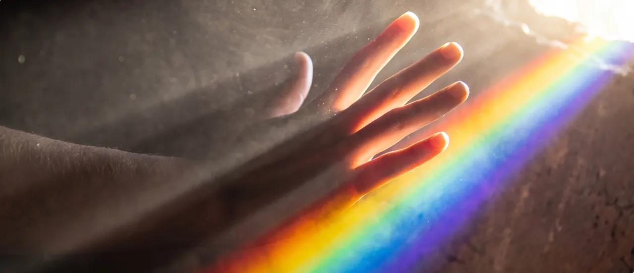 A hand reaching through a shaft of light with a rainbow prism spectrum cast across it