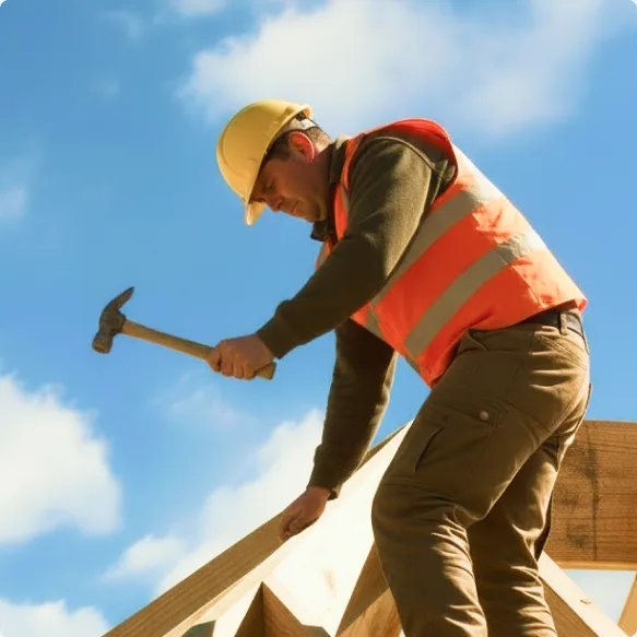 A roofer in a hard hat and high-visibility vest driving a nail into a roof truss against a blue sky
