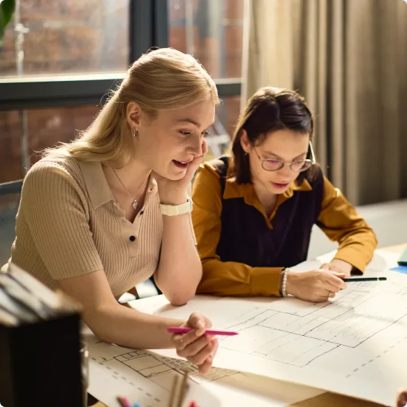 Two colleagues leaning over technical drawings at a desk by a window, marking them with pens